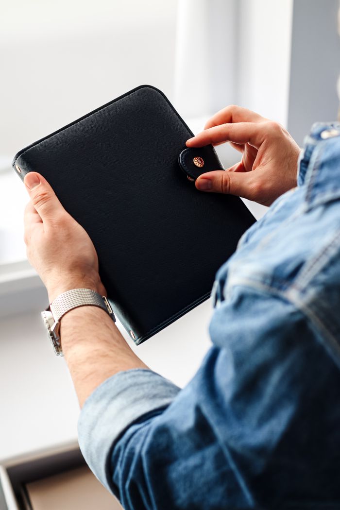 A person holding a sleek black leather portfolio with a button clasp near a window.